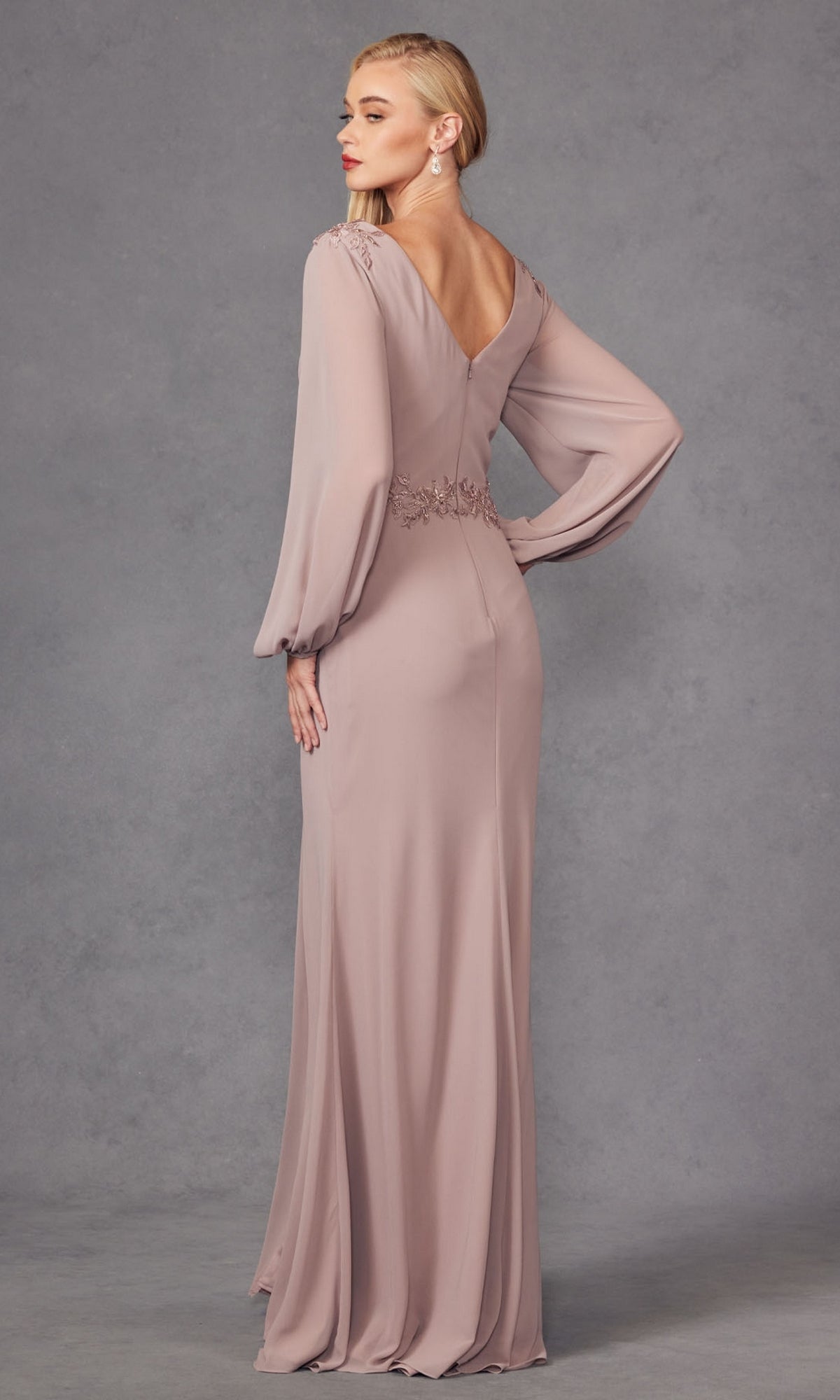 A woman faces away in the Juliet JTM16A V-Neck Long Sleeve Prom Gown, featuring sheer full-length sleeves and a V-shaped back, set against a plain gray background.